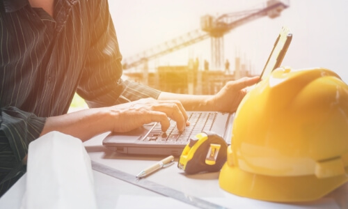 Woman typing on laptop with measuring tape and construction hat.
